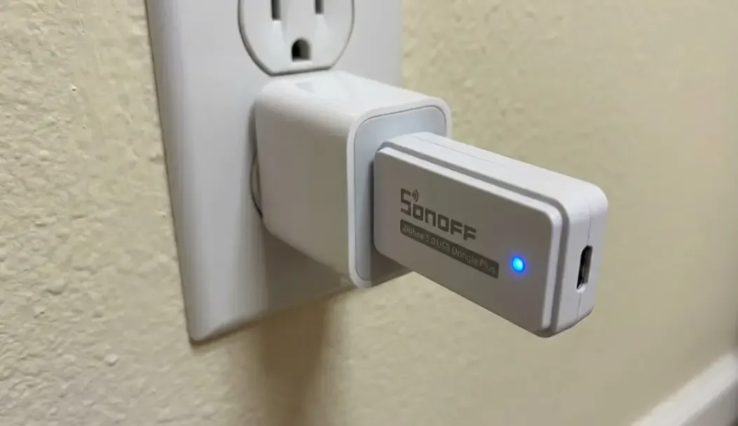 Primer plano de un repetidor Zigbee USB compacto Sonoff enchufado a un adaptador de corriente en la pared, mostrando el LED de estado indicando conexión exitosa a la red Mesh.