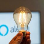 Mano sosteniendo una bombilla de filamento LED encendida con el logotipo de Amazon Alexa desenfocado al fondo, representando la iluminación inteligente.