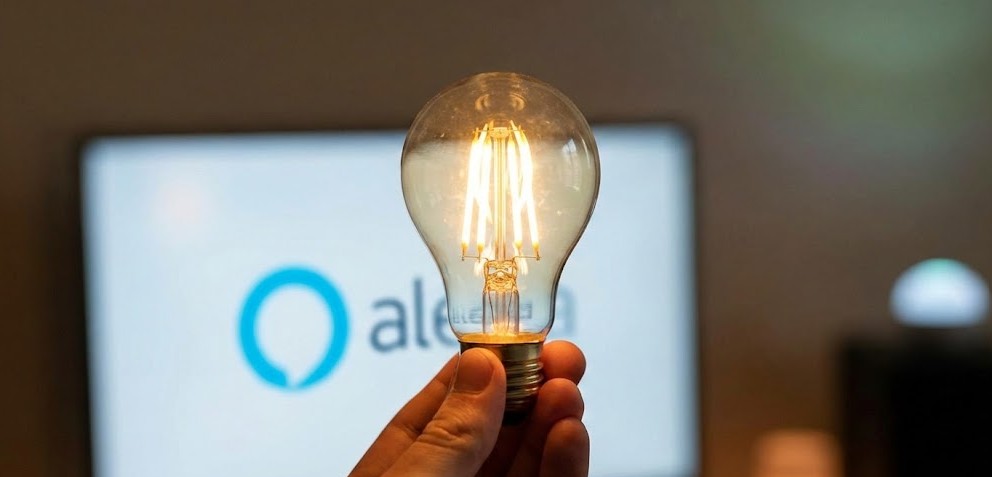 Mano sosteniendo una bombilla de filamento LED encendida con el logotipo de Amazon Alexa desenfocado al fondo, representando la iluminación inteligente.