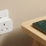 Primer plano de un enchufe inteligente blanco conectado a una toma de pared, junto a un teléfono móvil sobre una mesa de madera que muestra una gráfica verde de consumo de energía en su pantalla.