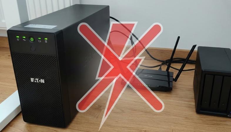 Sistema de Alimentación Ininterrumpida (SAI) negro en el suelo conectado a un router y un NAS, protegiendo contra cortes de luz. Opción B (SEO): Batería de respaldo SAI (UPS) tipo Eaton manteniendo encendidos el WiFi y el servidor durante un apagón.