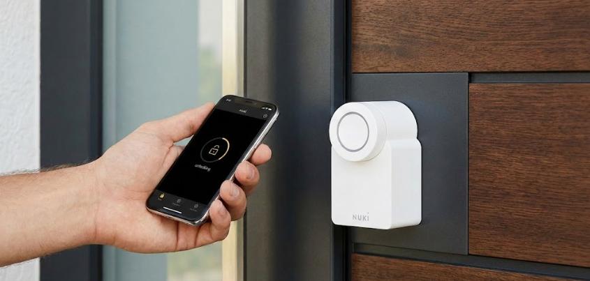 Primer plano de mano utilizando smartphone para desbloquear una cerradura inteligente Nuki blanca instalada en una puerta de entrada moderna de madera.