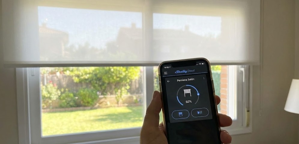 Mano sosteniendo un teléfono móvil con la aplicación Shelly Cloud abierta, mostrando una persiana ajustada al 50%, con la persiana física enrollable a medio bajar en la ventana del fondo.
