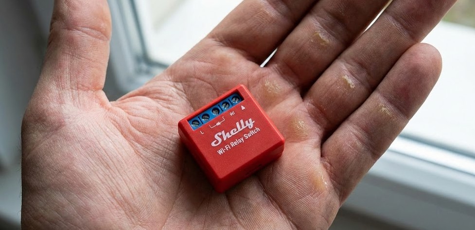 Relé inteligente Shelly de color rojo en la palma de una mano mostrando su tamaño compacto y diseño reducido.