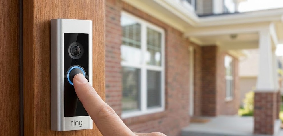 Dedo pulsando el botón azul iluminado de un timbre con video Ring montado en un marco de madera exterior.