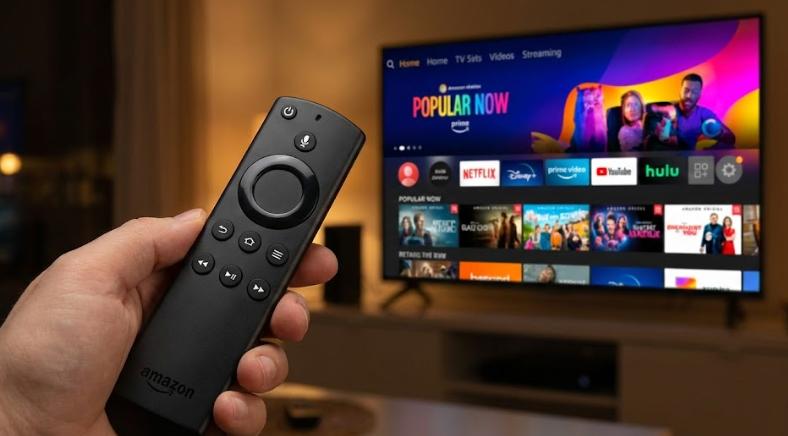 Primer plano de un mando a distancia Amazon Fire TV (Alexa Voice Remote) sostenido en la mano, con una Smart TV desenfocada al fondo mostrando una interfaz de aplicaciones de streaming.
