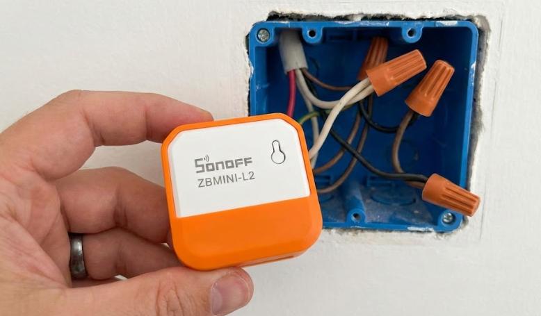 Mano sosteniendo el relé inteligente Sonoff ZBMINI-L2 (Extreme) frente a una caja de interruptor abierta, demostrando su tamaño compacto ideal para instalaciones sin cable neutro