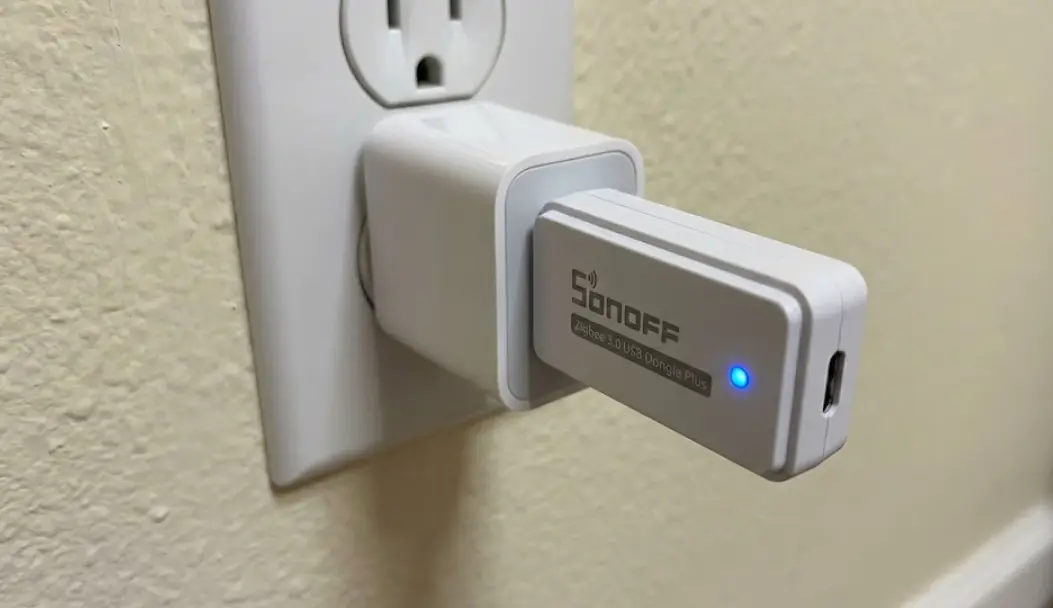 Primer plano de un repetidor Zigbee USB compacto Sonoff enchufado a un adaptador de corriente en la pared, mostrando el LED de estado indicando conexión exitosa a la red Mesh.